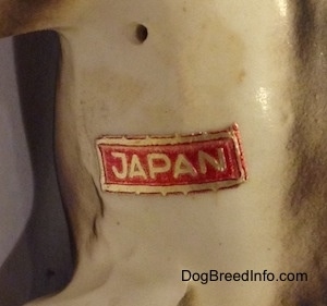 Close up - The underside of a black, brown and white ceramic Beagle figurine with a chain ID collar that reads "Beagle". There is a sticker on the figurine and the sticker reads - JAPAN.