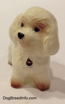 A white with tan miniature Bichon Frise dog figurine. The figurine has fine hair details. The dogs head has a round shape and the eyes are painted as black dots. There is a dog tag painted on the front. It has a black nose.