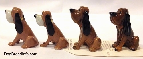 The left side of a line up of four Hagen-Renaker Bloodhound figurines. The dogs have very long hanging ears. Two of them have yellow collars.