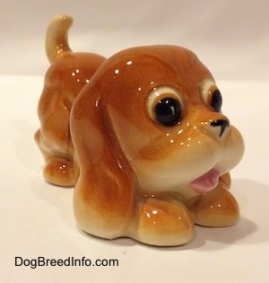 The front right side of a tan Cartoon style Bloodhound puppy figurine. The figurine is in a play bow pose.