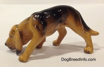 The left side of a brown and black Hagen Renaker Miniature Bloodhound figurine. The figurine has detailed arms and paws. It is stretching its front leg forward and its long tail is hanging down low.