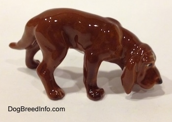 The right side of a Hagen-Renaker miniature red variation of a Bloodhound figurine. The figurine is very glossy. The dog has its nose to the ground.