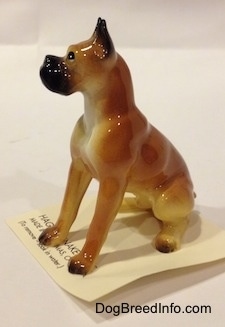 The front left side of a tan with black and white Boxer papa figurine. The figurine has black circles for eyes, but with a white dot inside it.