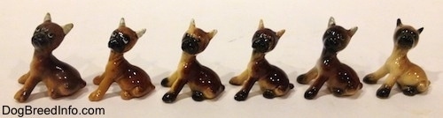 The left side of a line-up of the different color variations of a Boxer puppy figurine that is in a sitting pose. The heads of the figurines are turned forward.