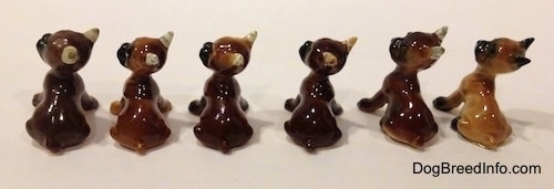 The back of a line-up of the different color variations of a Boxer puppy figurine that is in a sitting pose. The figurines are all glossy.