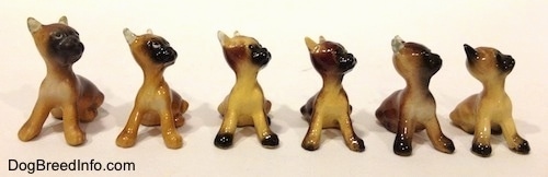 A line-up of the different color variations of a Boxer puppy figurine that is in a sitting pose. Four Sixths of the figurines have black paws.