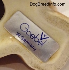 The underside of a brown with white Boxer puppy figurine that is in a sitting pose. There is a sticker on the bottom and on the sticker is a Goerbel W.Germany logo.
