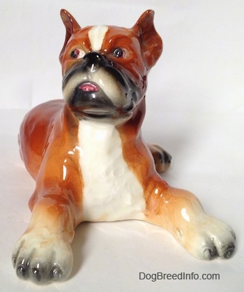 A fawn and white with black Boxer dog figurine in a laying pose. The figurine has its tongue painted to look like it is sticking out of its mouth. The dog has perk ears and a black nose.