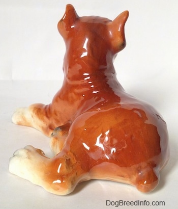 The back of a fawn and white with black Boxer dog figurine in a laying pose. The figurine is glossy.