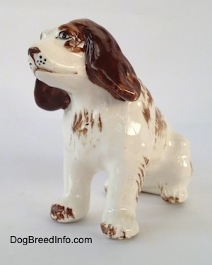 The front left side of a brown and white ceramic Brittany Spaniel that is in a sitting pose. The figurine has black whisker dots on its muzzle and detailed painted eyes.