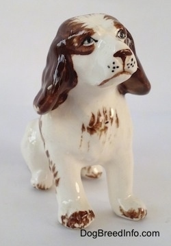 A ceramic brown and white Brittany Spaniel figurine that is in a sitting pose. The figurine has brown paw tips.