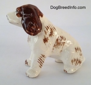 The left side of a brown and white ceramic Brittany Spaniel that is in a sitting pose. The figurine is glossy. The brown ears hang down to the sides.