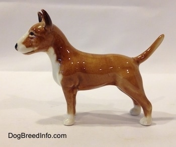 The left side of a brown with white Bull Terrier figurine. The paws of the figurine are white. The dog has perk ears and a tail that stands up. It has a black nose.