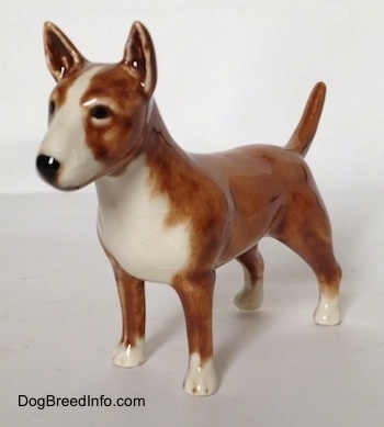 The front left side of a brown with white Bull Terrier figurine. The figurine has white paws and black painted eyes. Its tail is up in the air.