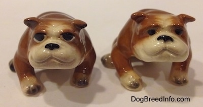 Two brown with white miniature Bulldogs that are in a sitting pose. The figurines have black circles for eyes.