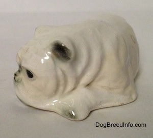 A miniature white English Bulldog figurine that is in a lying down pose with a brown spot over its left eye. The ears of the figurines are hard to tell the difference between its body.