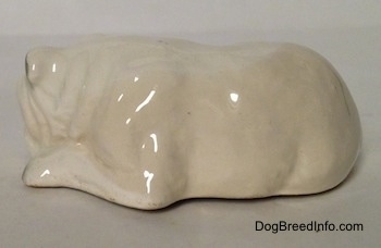 The left side of a miniature white English Bulldog figurine that is in a lying down pose with a brown spot over its left eye. The arms of the figurine are attached to its body.