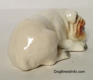 The back right side of a miniature white English Bulldog figurine that is in a lying down pose with a brown spot over its left eye. The figurine is very glossy.