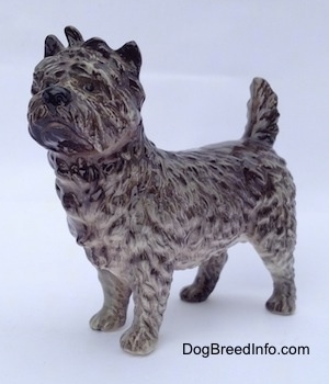 The front left side of a gray and white Cairn Terrier figurine. The figurine has black circles for eyes, a wavy textured coat, a tail that is up in the air and small perk ears.