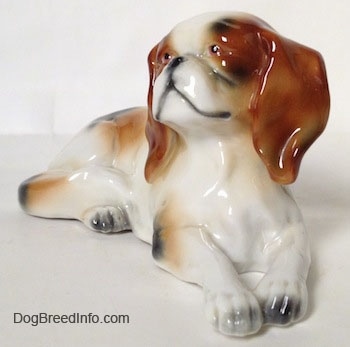A white with brown and black Cavalier King Charles Spaniel figurine that is laying down. The figurine has black at the tips of its paws.