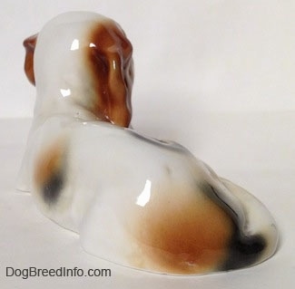 The back left side of a white with brown and black Cavalier King Charles Spaniel figurine that is laying down. The figurine is glossy.