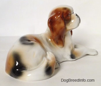 The back right side of a white with brown and black Cavalier King Charles Spaniel figurine that is laying down. The tail of the figurine is hard to differentiate from the body.