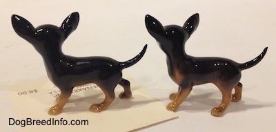 The left side of two slightly different black with tan Chihuahua figurines. The figurines have long tails.