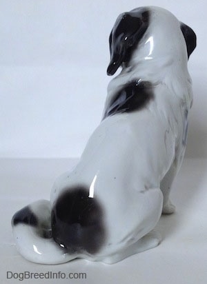 The back of white with black Japanese Chin dog figurine. The tail of the figurine is long and around the left leg of the figurine.