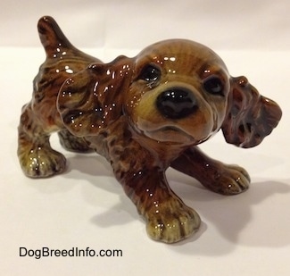 A brown and tan Cocker Spaniel puppy figurine is standing across a beige backdrop and it is looking forward.
