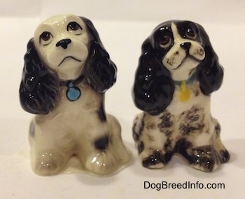 Two different ceramic Cocker Spaniel figurines. Are looking up and there heads are tilted to the left. The figurine on the left has light eye details and the right figurine has fine eye details. The dogs have black noses and black lips. One dog has a blue dog tag and the other has a yellow dog tag.
