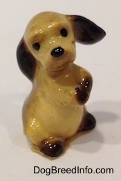 The front right side of a tan with brown ceramic Cocker Spaniel puppy figurine that is sitting on its hind legs and its front paws are together. Its head is tilted to the left and its ears are swinging to the right.