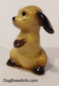 The left side of a tan with brown ceramic Cocker Spaniel puppy figurine that is sitting on its hind legs and its front paws are together. The figurine is very glossy.