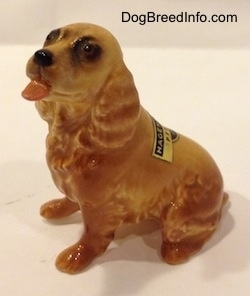 The left side of a tan ceramic Cocker Spaniel figurine that is sticking its tongue out. There is a tag on the back of the figurine.