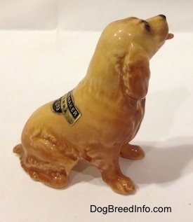The right side of a tan ceramic Cocker Spaniel figurine that is sticking its tongue out. There is a tag on the back of the figurine and it has fine hair details.