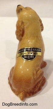 The back of a tan ceramic Cocker Spaniel figurine that is sticking its tongue out. There is a tag on its back that reads - Hagen-Renaker PIP EMMA 19©53