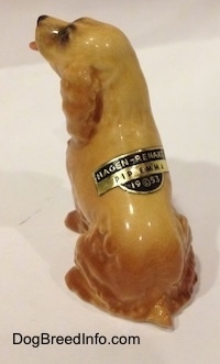 The back left side of a tan ceramic Cocker Spaniel figurine that is sticking its tongue out. There is a tag on its back that reads - Hagen-Renaker PIP EMMA 19©53. The figurine has hair details.
