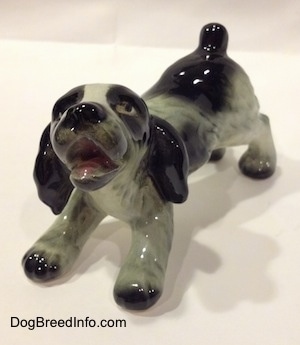The front left side of a white and black Cocker Spaniel puppy figurine. The figurine is looking up and its mouth is open.