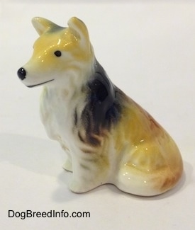The left side of a tan, black and white rough coated Collie dog figurine in a sitting pose. The figurine has black circles for eyes.