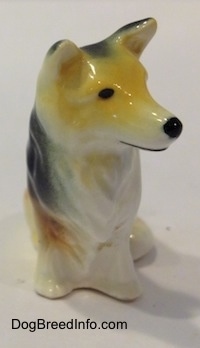 A figurine of a tan, black and white rough coated Collie dog that is in a sitting pose. The back of the figurines ears are black.