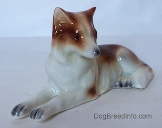 The front left side of figurine that is a brown and white Collie dog in a lying down pose. The figurine has few details in its paws. The nails on the paws are black.