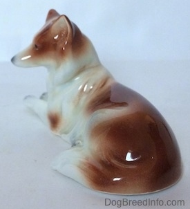 The back right side of a figurine that is a brown and white Collie dog in a lying down pose.