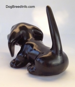 The back left side of a black Dachshund figurine. The figurine has a long body and its tall tail is in the air.