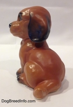 The back left side of a brown with black Dachshund puppy in a sitting pose figurine. The figurines tail is medium length relative tot he size of the figurine.