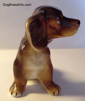 A ceramic brown with tan Dachshund figurine. The figurine has big ears and a white chest. The figurine has big paws with black nails on it.