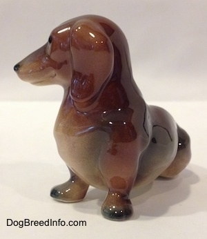 The front left side of a brown and black Dachshund figurine in a sitting pose. The figurine has black tipped paws.