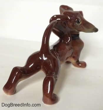 The back right side of a brown with white Dachshund figurine in a stretching pose. The figurine has on a black collar.