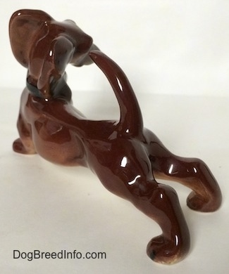 The back left side of a figurine that is a brown with white Dachshund figurine in a strecthing pose. The figurine has a long arching tail.