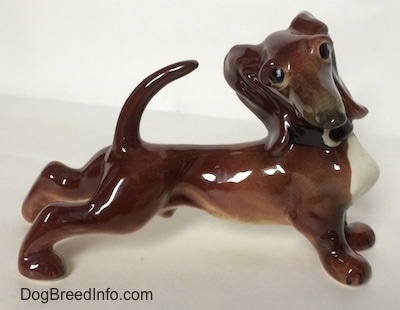 The right side of a brown with white Dachshund figurine that is in a strecthing pose. The figurine has a long body and it is glossy.