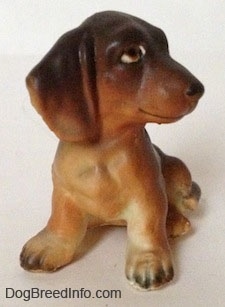 A brown with tan Dachshund puppy in a sitting pose figurine. The figurine has a detailed body, it has big paws with black nails.