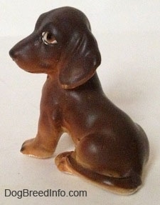 The back left side of a brown with tan Dachshund puppy in a sitting pose figurine. The figurine has a long body and it has a long tail across its leg.